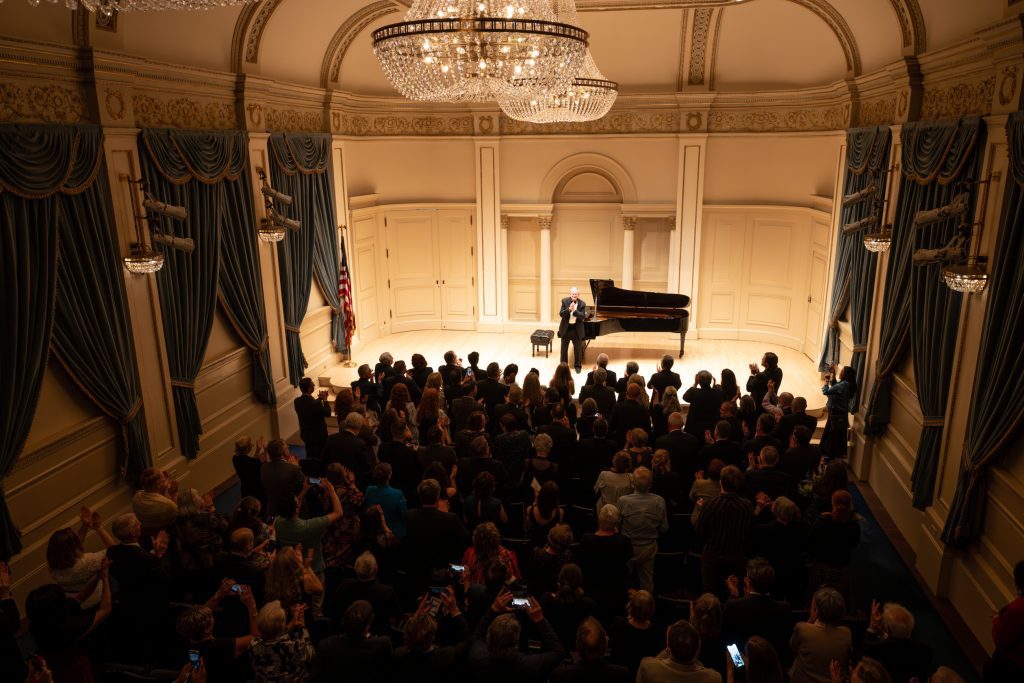 Marvin Blickenstaff stands at center stage in front of a grand piano at Carnegie Hall’s Weill Recital Hall, addressing a full audience giving a standing ovation.