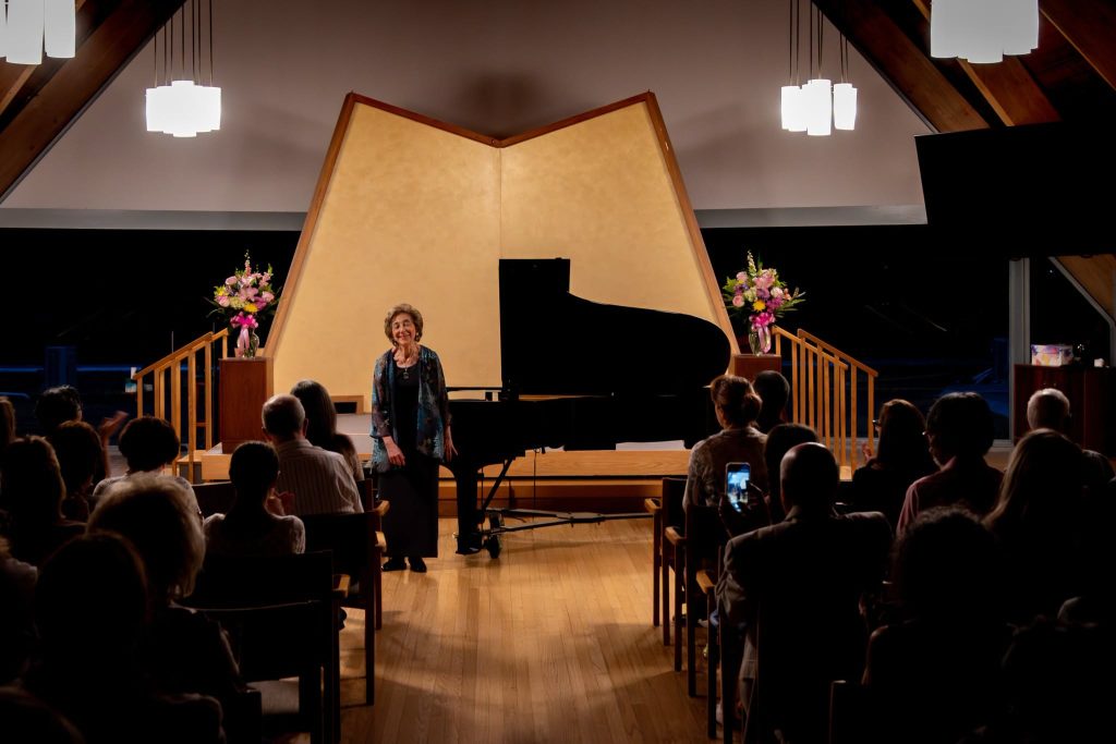 Phyllis Lehrer is acknowledged by an audience in a recital space.