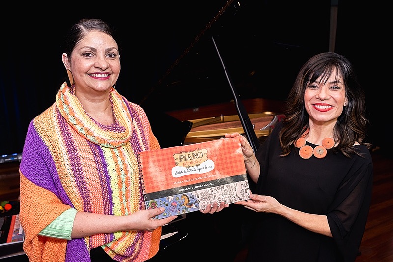 A photograph of two women holding a sheet music book in front of a grand piano.